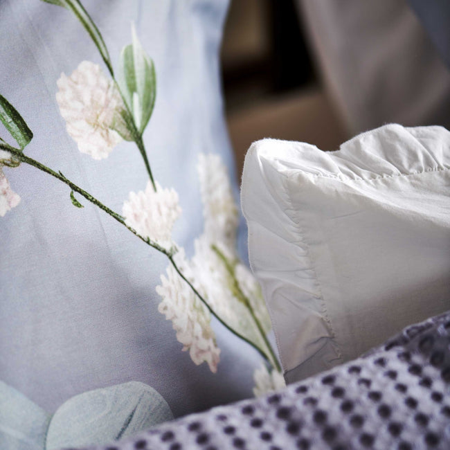 Fleur Festive lavender blue dekbedovertrek heeft hele grote bloemen in lichte pasteltinten in blauw, roze en oranje schakeringen op een blauw / paarse achtergrond. De achterkant van het dekbedovertrek is effen beige. De kussens hebben aan beide kanten een paar bloemen en een bloesemtak.