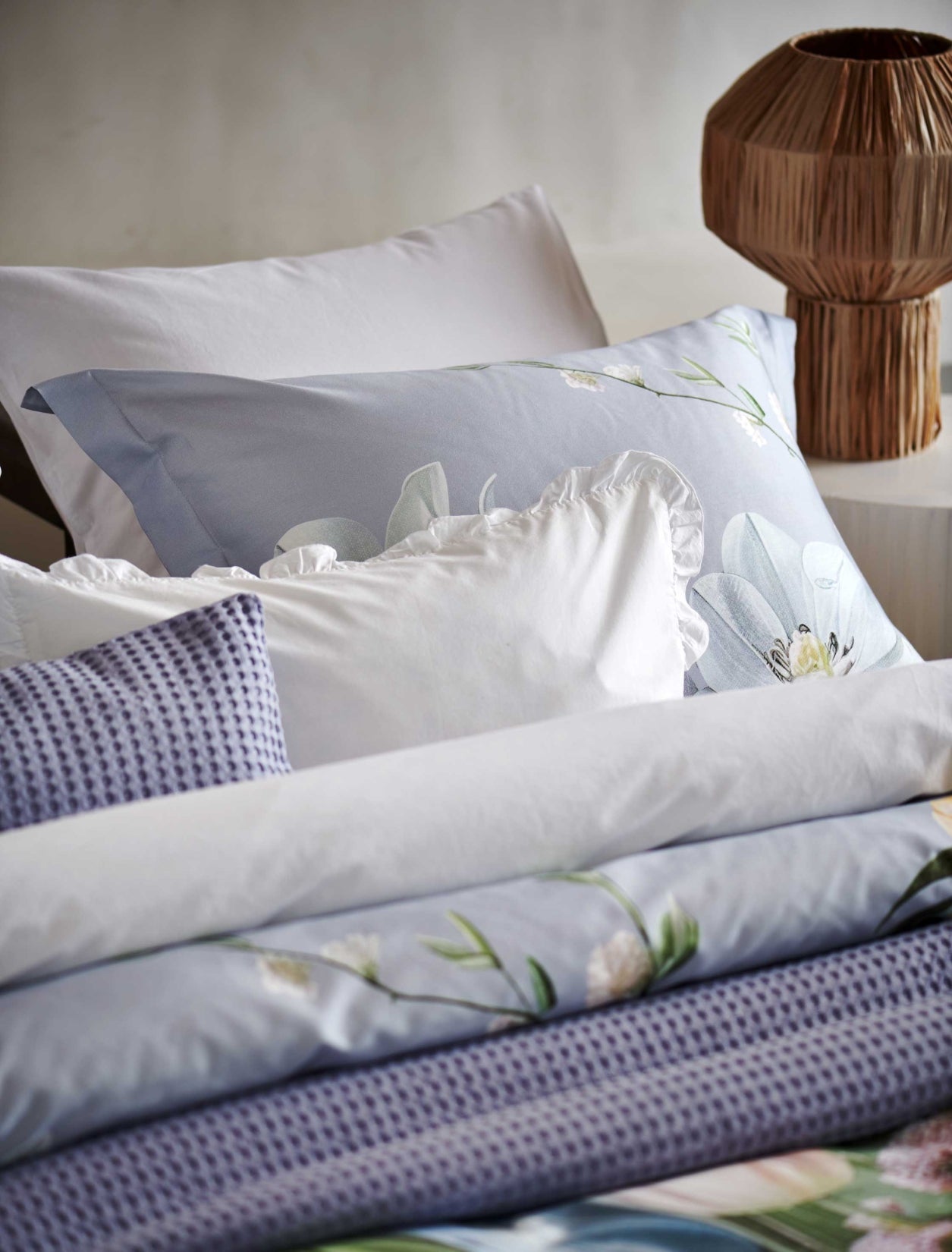 Fleur Festive lavender blue dekbedovertrek heeft hele grote bloemen in lichte pasteltinten in blauw, roze en oranje schakeringen op een blauw / paarse achtergrond. De achterkant van het dekbedovertrek is effen beige. De kussens hebben aan beide kanten een paar bloemen en een bloesemtak.
