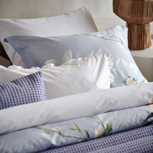 Fleur Festive lavender blue dekbedovertrek heeft hele grote bloemen in lichte pasteltinten in blauw, roze en oranje schakeringen op een blauw / paarse achtergrond. De achterkant van het dekbedovertrek is effen beige. De kussens hebben aan beide kanten een paar bloemen en een bloesemtak.