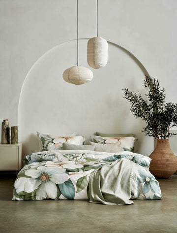 Een bed met een beige dekbedovertrek met daarop groene bladeren en bloemen. Boven het bed hangen lampen en er is een groen plaid over het bed heen gedrapeert