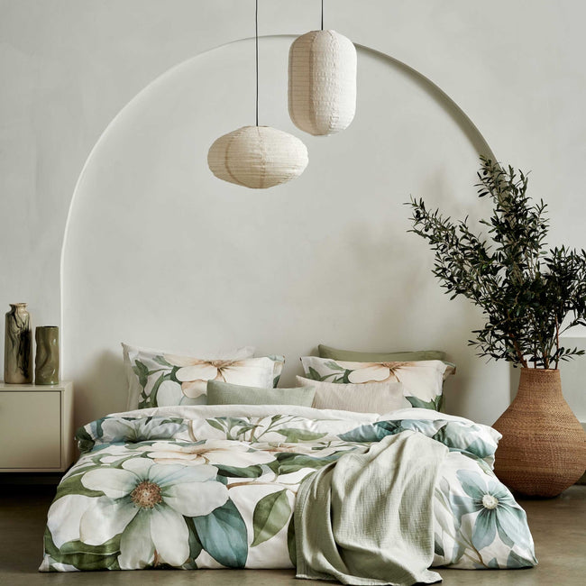 Een bed met een beige dekbedovertrek met daarop groene bladeren en bloemen. Boven het bed hangen lampen en er is een groen plaid over het bed heen gedrapeert