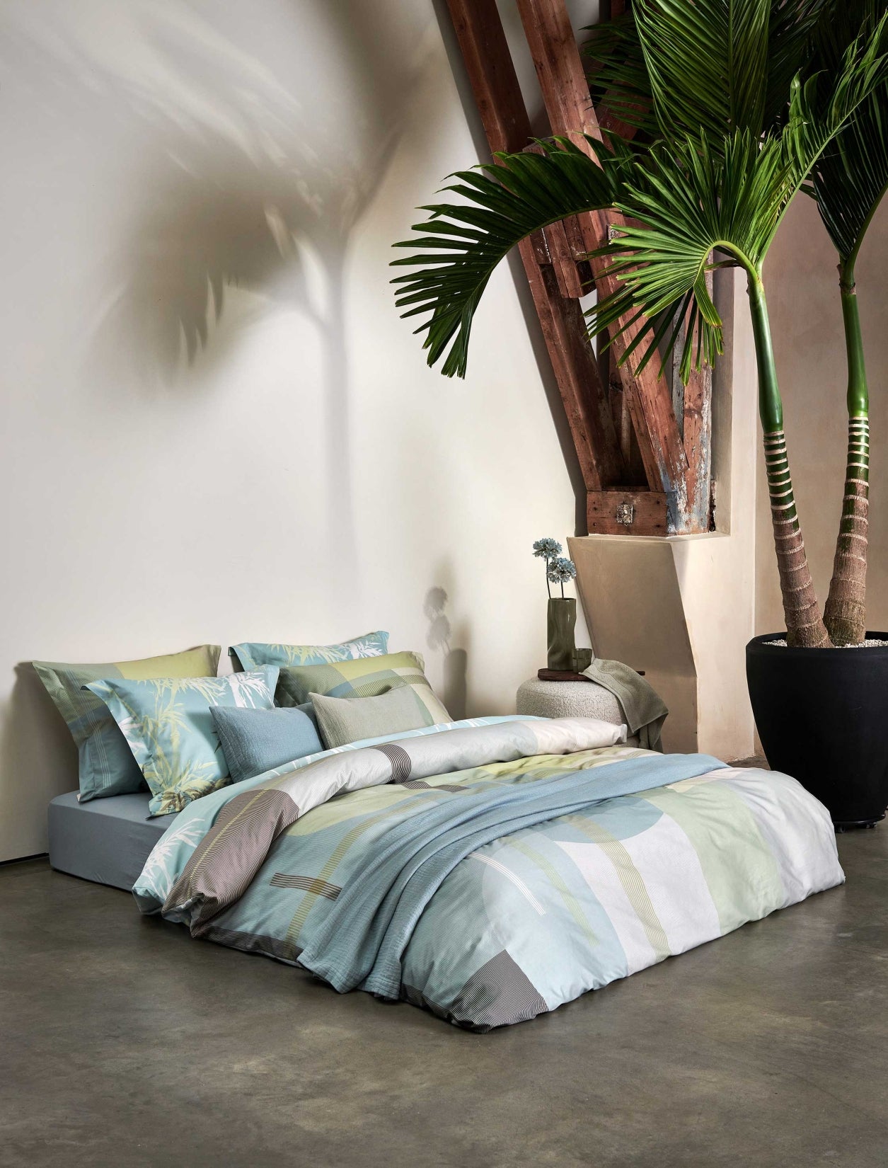 een bed in een rustige omgeving met een plant op de achtergrond. Het bed heeft een dekbedovertrek met groene en blauwe kleuren in een grafische print en een dekbedovertrek met palmbomen in groene en blauwe kleuren