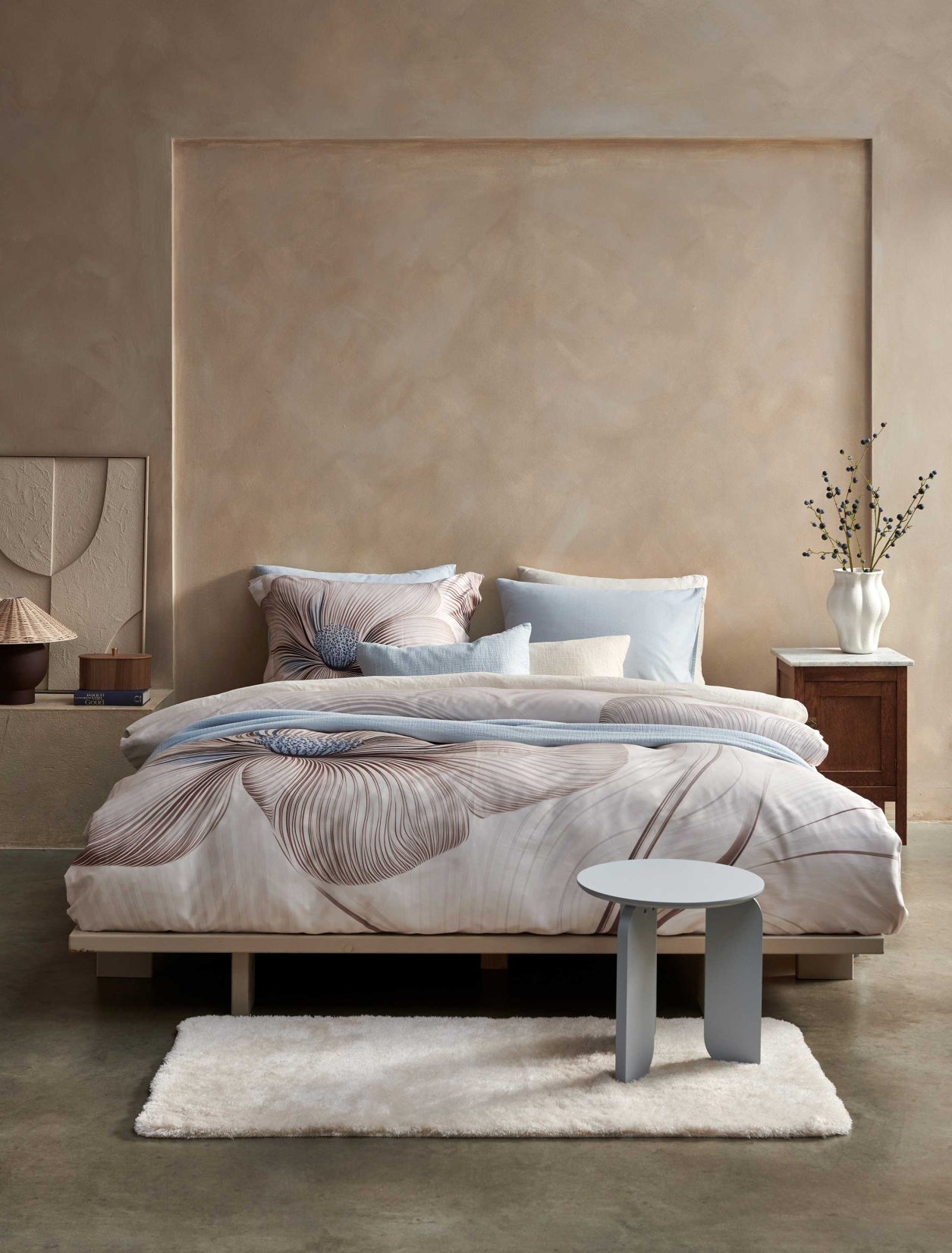 Een bed in een rustige omgeving met een beige dekbedovertrek set met een grote bloem in bruine en blauwe kleuren. Op het bed ligt een lichtblauw plaid