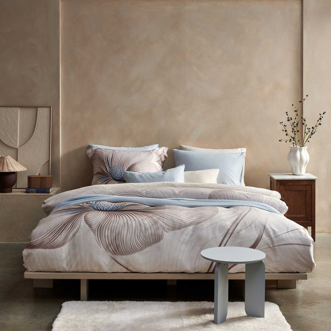 Een bed in een rustige omgeving met een beige dekbedovertrek set met een grote bloem in bruine en blauwe kleuren. Op het bed ligt een lichtblauw plaid