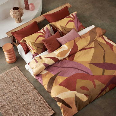 Sunset Nights multi dekbedovertrek heeft verschillende soorten bladeren in een abstracte tekenstijl in bruin, roze en rood. De achterkant van het dekbedovertrek is effen beige. De kussens hebben een kleinere versie van de bladeren.