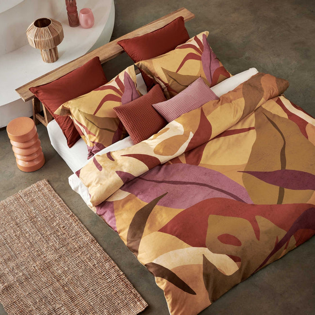 Sunset Nights multi dekbedovertrek heeft verschillende soorten bladeren in een abstracte tekenstijl in bruin, roze en rood. De achterkant van het dekbedovertrek is effen beige. De kussens hebben een kleinere versie van de bladeren.
