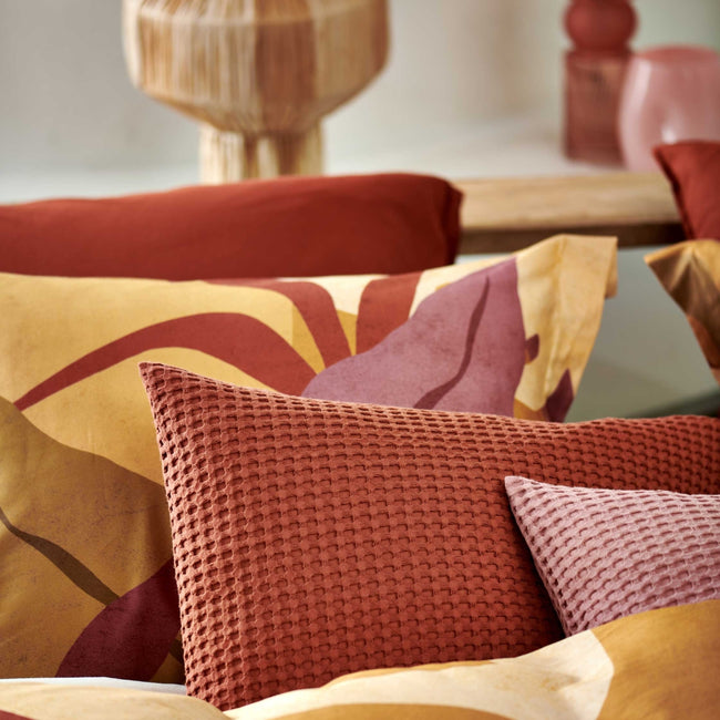 Sunset Nights multi dekbedovertrek heeft verschillende soorten bladeren in een abstracte tekenstijl in bruin, roze en rood. De achterkant van het dekbedovertrek is effen beige. De kussens hebben een kleinere versie van de bladeren.
