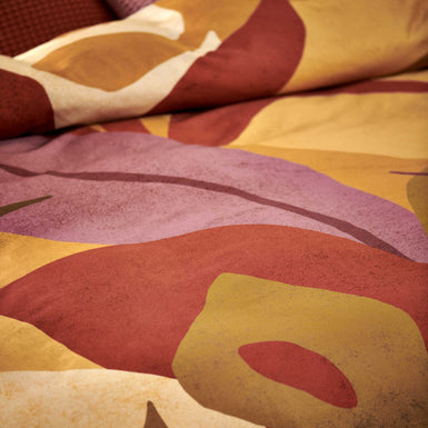 Sunset Nights multi dekbedovertrek heeft verschillende soorten bladeren in een abstracte tekenstijl in bruin, roze en rood. De achterkant van het dekbedovertrek is effen beige. De kussens hebben een kleinere versie van de bladeren.