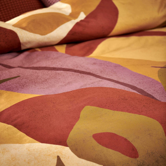 Sunset Nights multi dekbedovertrek heeft verschillende soorten bladeren in een abstracte tekenstijl in bruin, roze en rood. De achterkant van het dekbedovertrek is effen beige. De kussens hebben een kleinere versie van de bladeren.