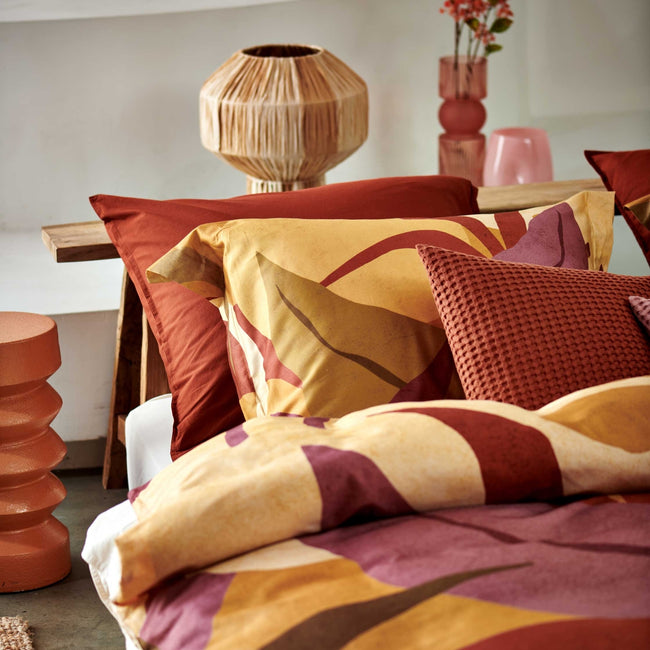 Sunset Nights multi dekbedovertrek heeft verschillende soorten bladeren in een abstracte tekenstijl in bruin, roze en rood. De achterkant van het dekbedovertrek is effen beige. De kussens hebben een kleinere versie van de bladeren.