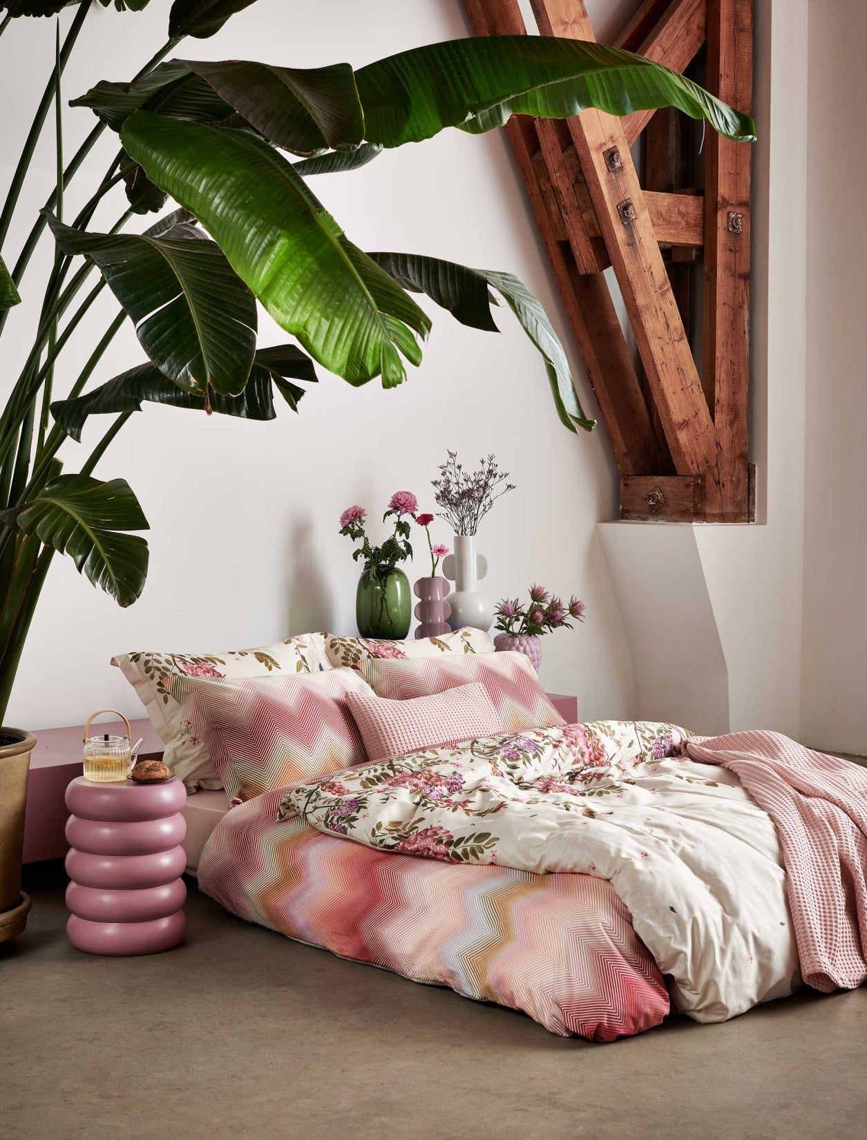 Een bed met een roze zigzag dekbedovertrek en een dekbedovertrek met roze en paarse bloemen. Rond het bed staan prachtige planten en roze bloemen