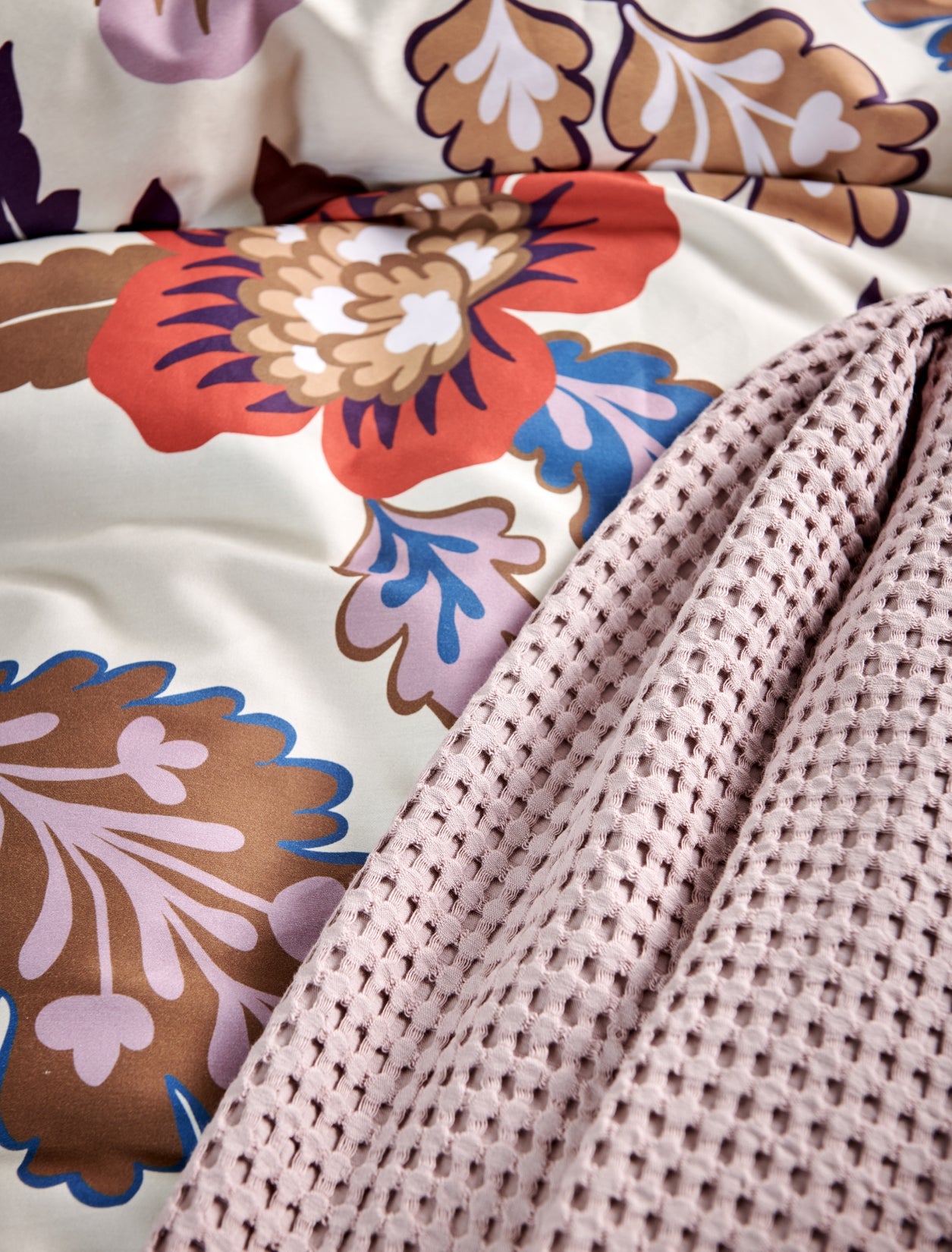 detail shot van een dekbedovertrek met rode bloemen en blauwe en paarse kleuren en een plaid in een lila kleur