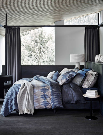 Een bed in een rustige omgeving met een dekbedovertrek met grafische vormen in donkerblauwe kleuren en een grijs plaid