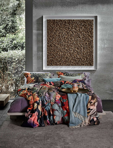 een bed in een rustige omgeving met een dekbedovertrek met een print in oranje, paarse en blauwe kleuren en een plaid van wol mix in een blauwe kleur