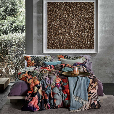 een bed in een rustige omgeving met een dekbedovertrek met een print in oranje, paarse en blauwe kleuren en een plaid van wol mix in een blauwe kleur