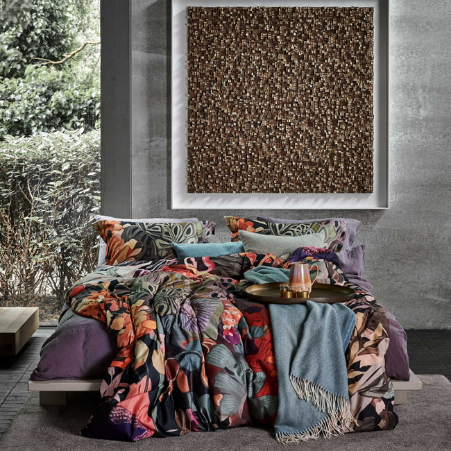 een bed in een rustige omgeving met een dekbedovertrek met een print in oranje, paarse en blauwe kleuren en een plaid van wol mix in een blauwe kleur