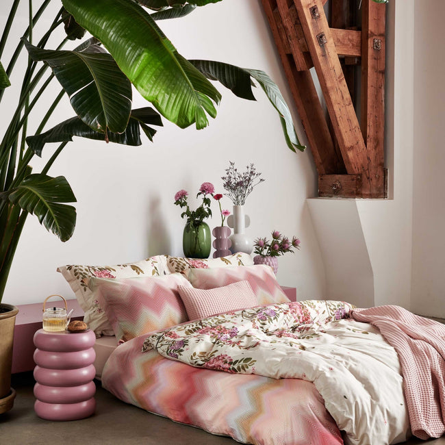 Rose Acacia cream tan dekbedovertrek heeft aan de bovenkant roze en paarse bloemen, die aan takken hangen met kleine dwarrelende blaadjes. De achterkant van het dekbedovertrek is effen beige. Het design oogt rustig en zacht.
