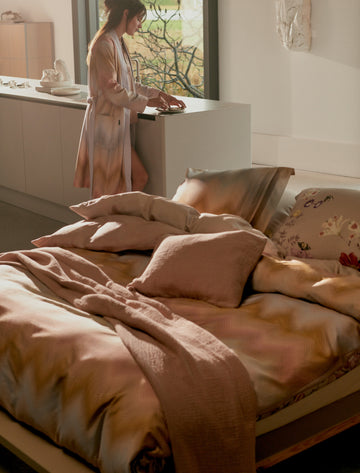 een vrouw die aan het keuken aanrecht staat met een badjas aan en een bed met een dekbedovertrek in paarse tinten